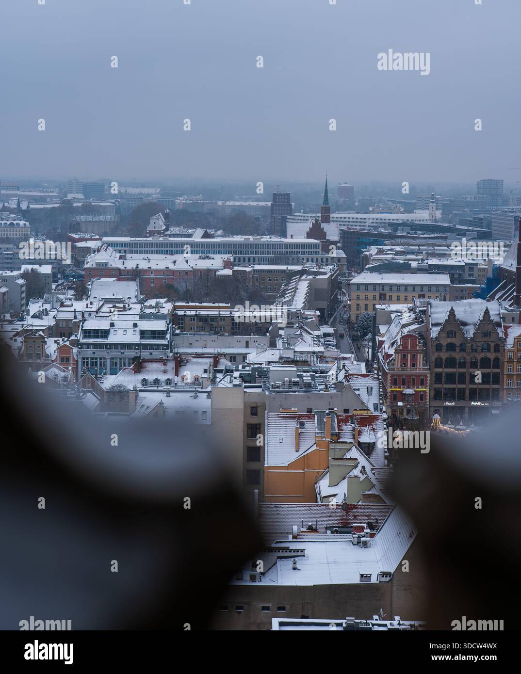I tetti della città Vecchia di Wroclaw sono ricoperti di polvere di neve, mentre una guglia di mattoni sorge vicino a edifici moderni. La morbida foschia e la luce soffusa suggeriscono un tranquillo pomeriggio invernale Foto Stock