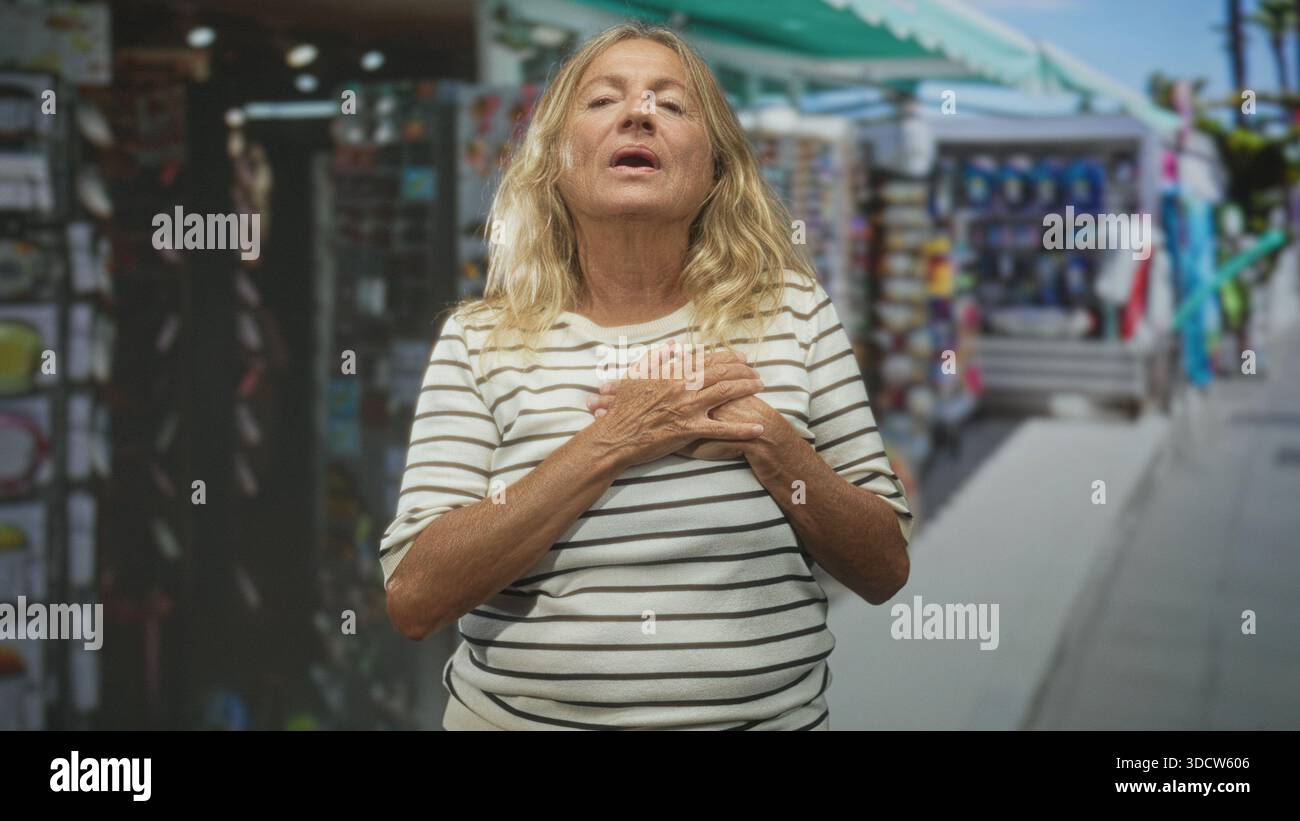 Donna anziana che stringe le mani al petto e inclina la testa all'indietro a occhi chiusi su una strada costeggiata da bancarelle del mercato; riflesso di gratitudine. Foto Stock