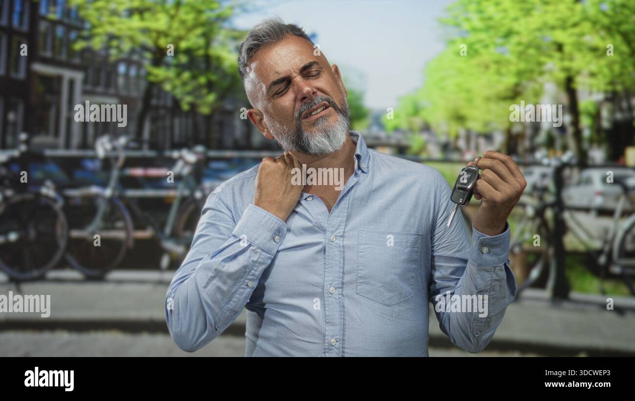 Uomo che tiene in mano la chiave della macchina con la mano sul collo per dolore al collo per strada ad amsterdam, accanto al canale; disagio per gli effetti personali persi. Foto Stock