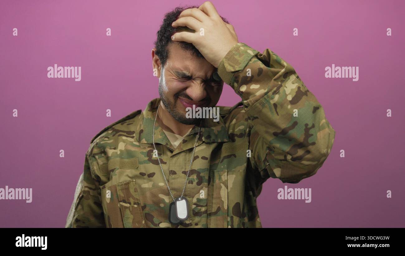 Il giovane soldato ispanico in uniforme mimetica tiene in mano le targhette per cani d'argento e preme mano sulla fronte in studio rosa; stress. Foto Stock