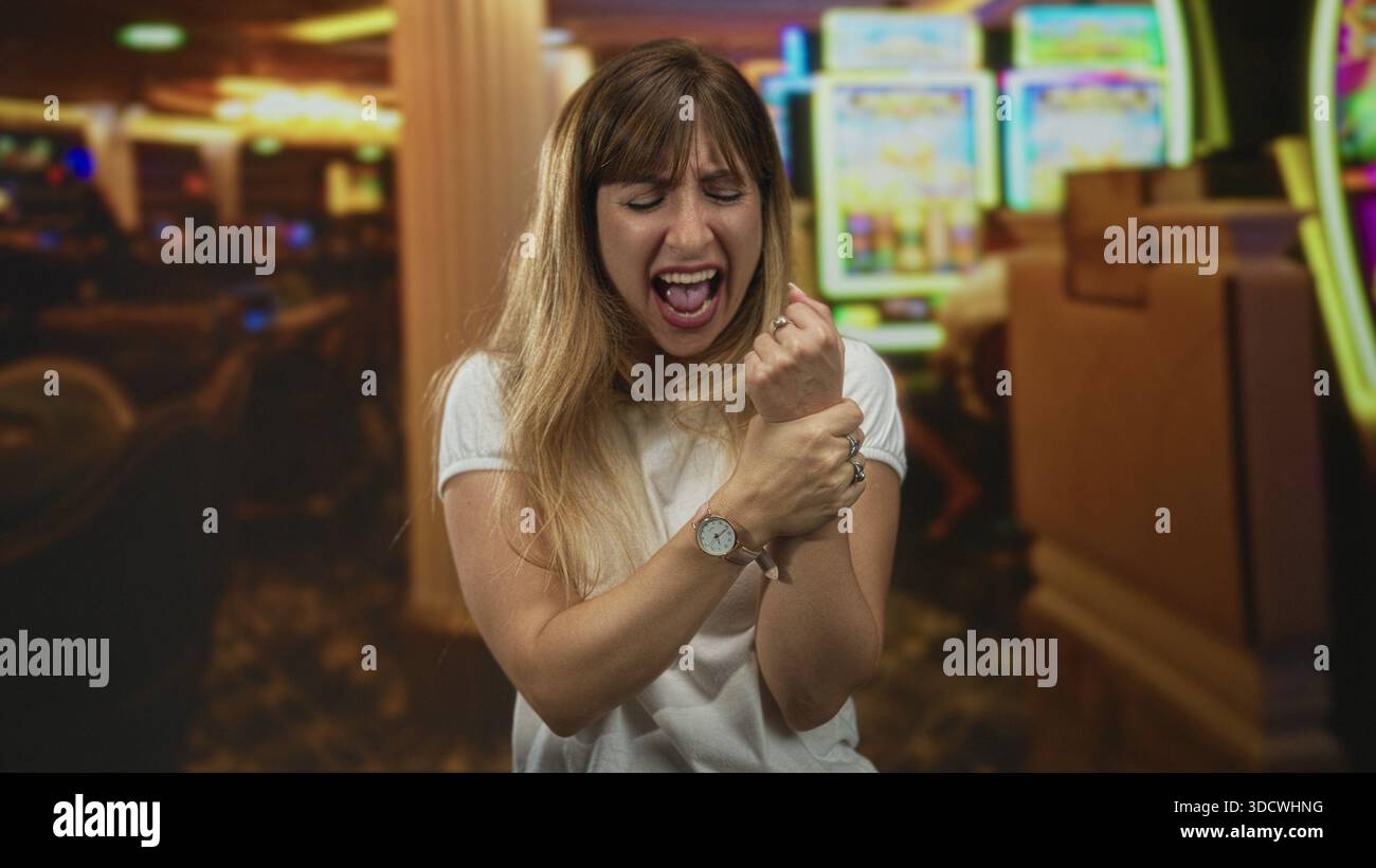 Donna che stringe il polso e urla contro una slot machine all'interno di un casinò, occhi chiusi e tesi; frustrazione. Foto Stock