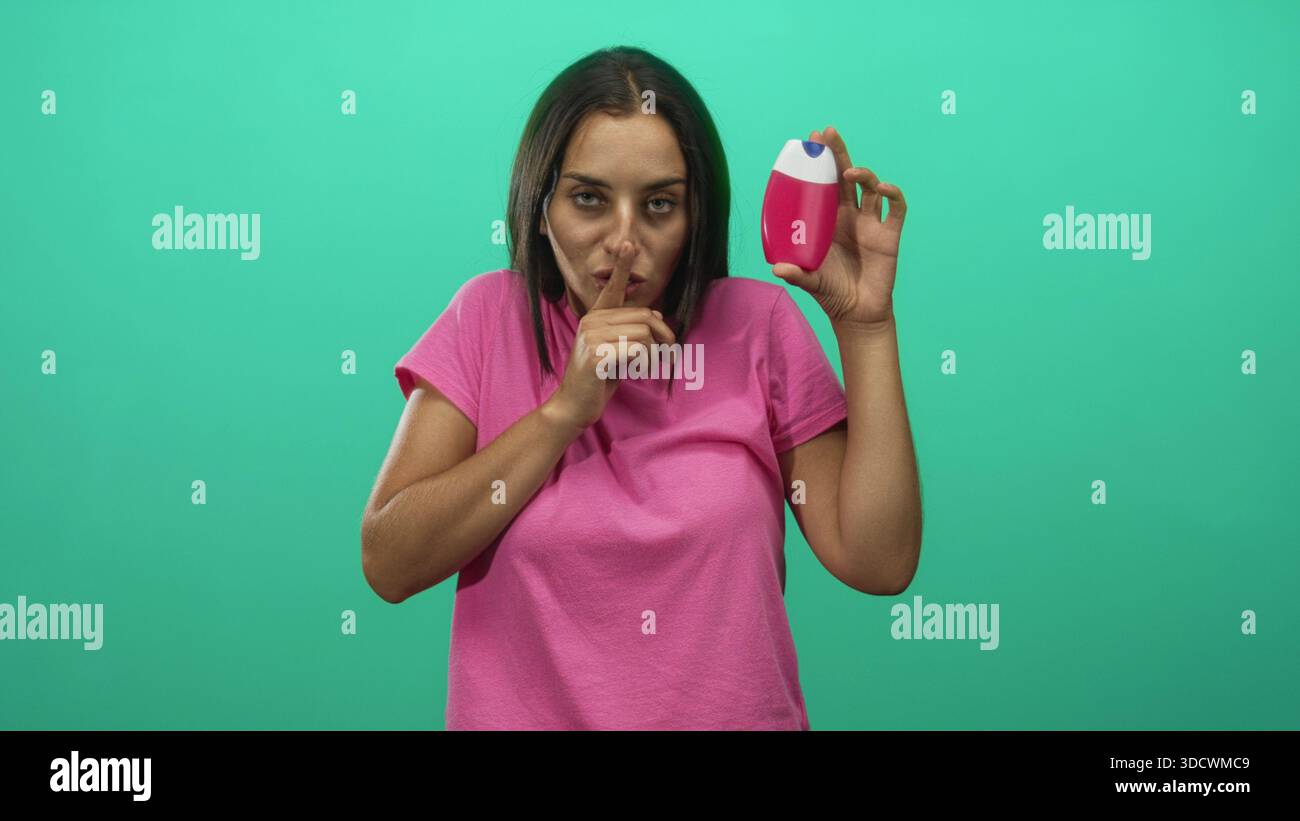 Donna che tiene una bottiglia di deodorante rosa vicino al viso, dita alle labbra per il silenzio in uno studio verde; disgustosa esitazione segreta. Foto Stock