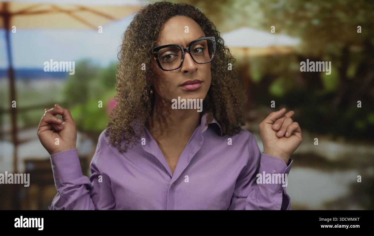 Donna con capelli ricci e occhiali punta entrambe le dita indice lateralmente nel parco con ombrelli e fogliame; indecisione. Foto Stock