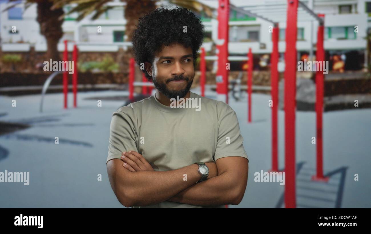 L'uomo con le braccia incrociate si trova in una palestra all'aperto circondata da attrezzature da palestra rosse, adagiata su uno sfondo urbano moderno con palme. Foto Stock