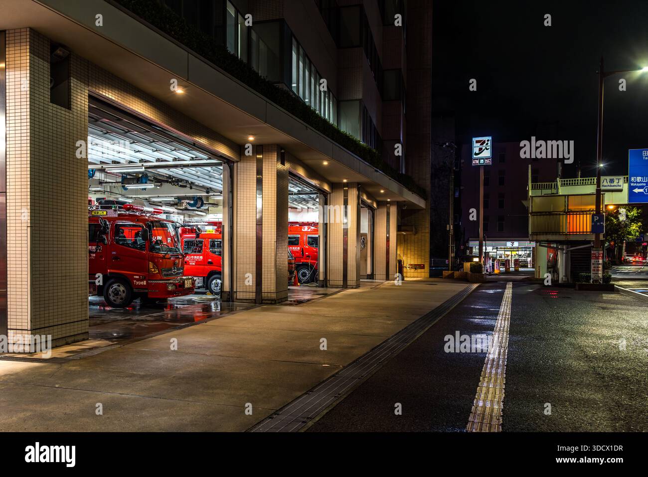 I camion dei pompieri rossi del corpo dei vigili del fuoco di Wakayama si trovano in un garage a Wakayama, Giappone, con le porte aperte sulla strada. L'asfalto bagnato dalla pioggia riflette la luce del garage. I camion portano la scritta "Wakayama City Fire Bureau" (和歌山市消防局). Foto Stock