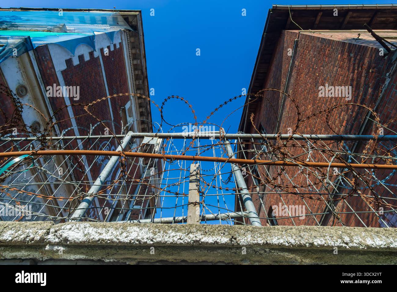 Riprese grandangolari ad angolo basso di edifici industriali abbandonati viste attraverso una recinzione di filo spinato arrugginito. Foto Stock