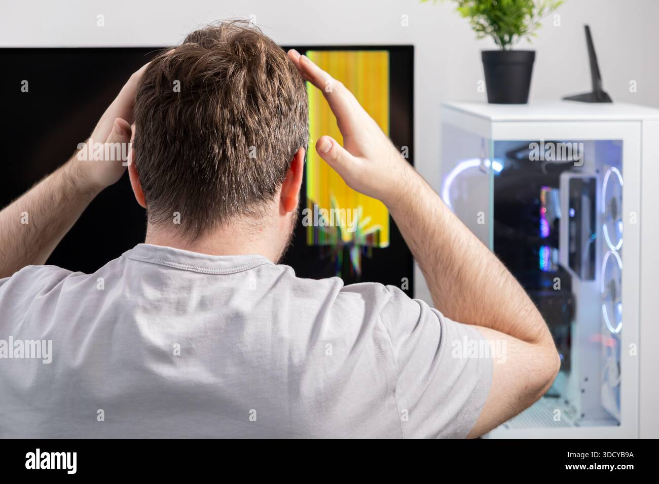 Vista posteriore di un uomo in difficoltà che afferra la testa, fissa uno schermo rotto con un intenso glitch giallo, che enfatizza i guasti hardware e u Foto Stock