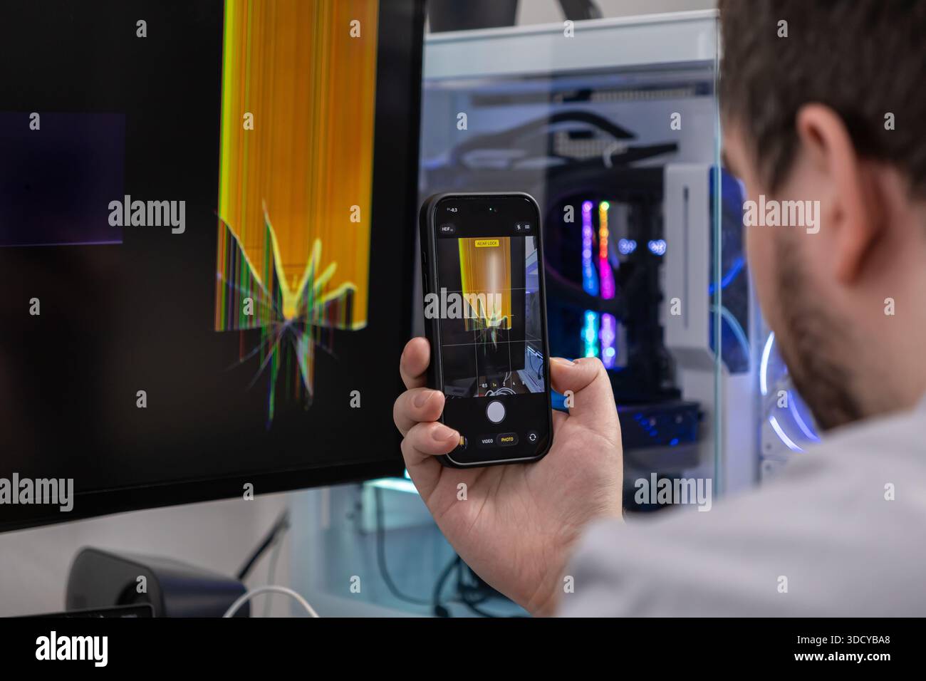 Primo piano di un uomo con uno smartphone, che documenta il monitor di un computer desktop danneggiato con uno schermo incrinato e un display distorto per la risoluzione dei problemi Foto Stock