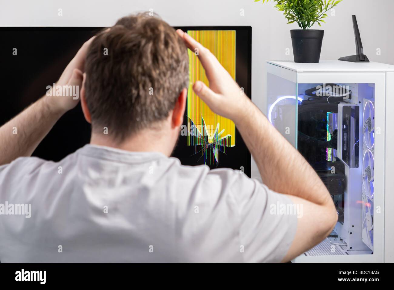 Vista posteriore di un uomo con le mani sulla testa, che esprime disagio guardando uno schermo del computer gravemente rotto che mostra difetti colorati e un moderno PC setu Foto Stock