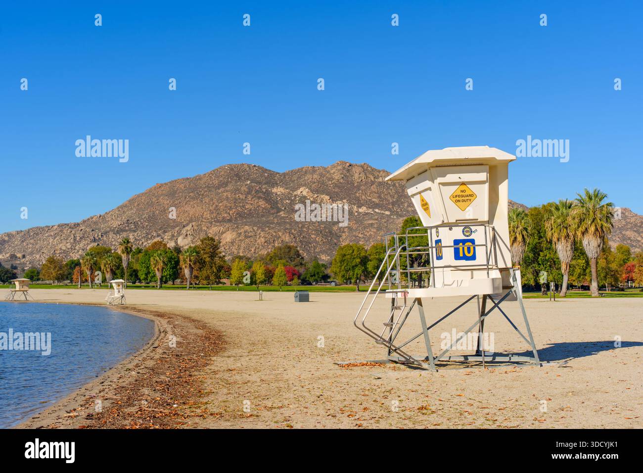 Torre bagnino sulla spiaggia del lago Perris con foglie autunnali, acque tranquille del lago, montagne rocciose e alberi colorati sotto un cielo azzurro. Foto Stock