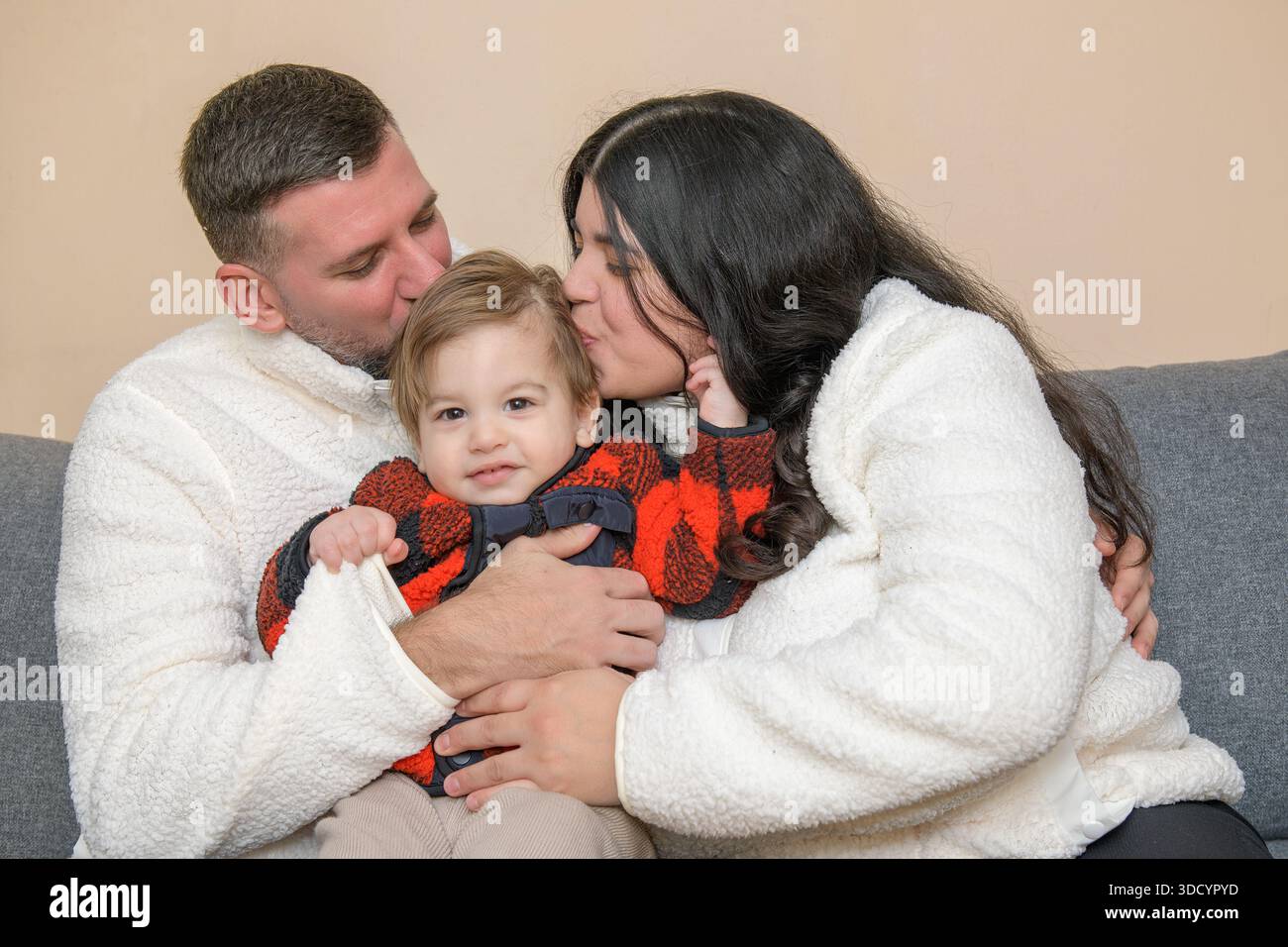 I genitori baciano il loro bambino seduto insieme su un divano al chiuso. Foto Stock
