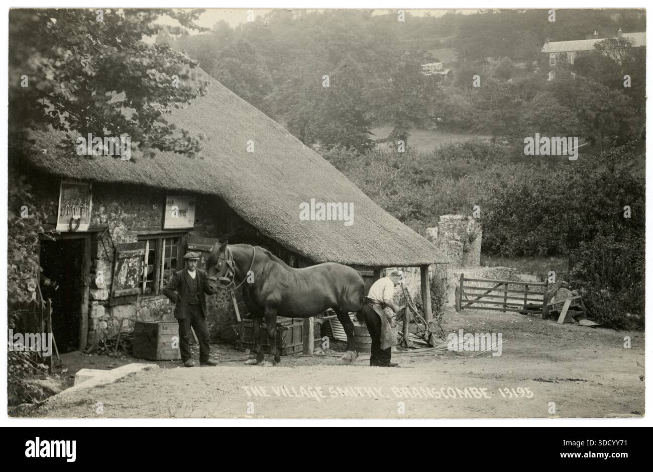Cartolina originale dei primi anni '1900 di affascinante scena rurale - un contadino o una mano di fattoria tiene il suo cavallo pesante / funzionante mentre viene spinto dal fabbro / contadino - fuori Branscombe Village Smithy / Forge, Devon, Inghilterra, fabbro vittoriano del Regno Unito. Questa fucina del XVIII secolo è la più antica fucina di paglia funzionante in Inghilterra. Circa 1920 anni Foto Stock