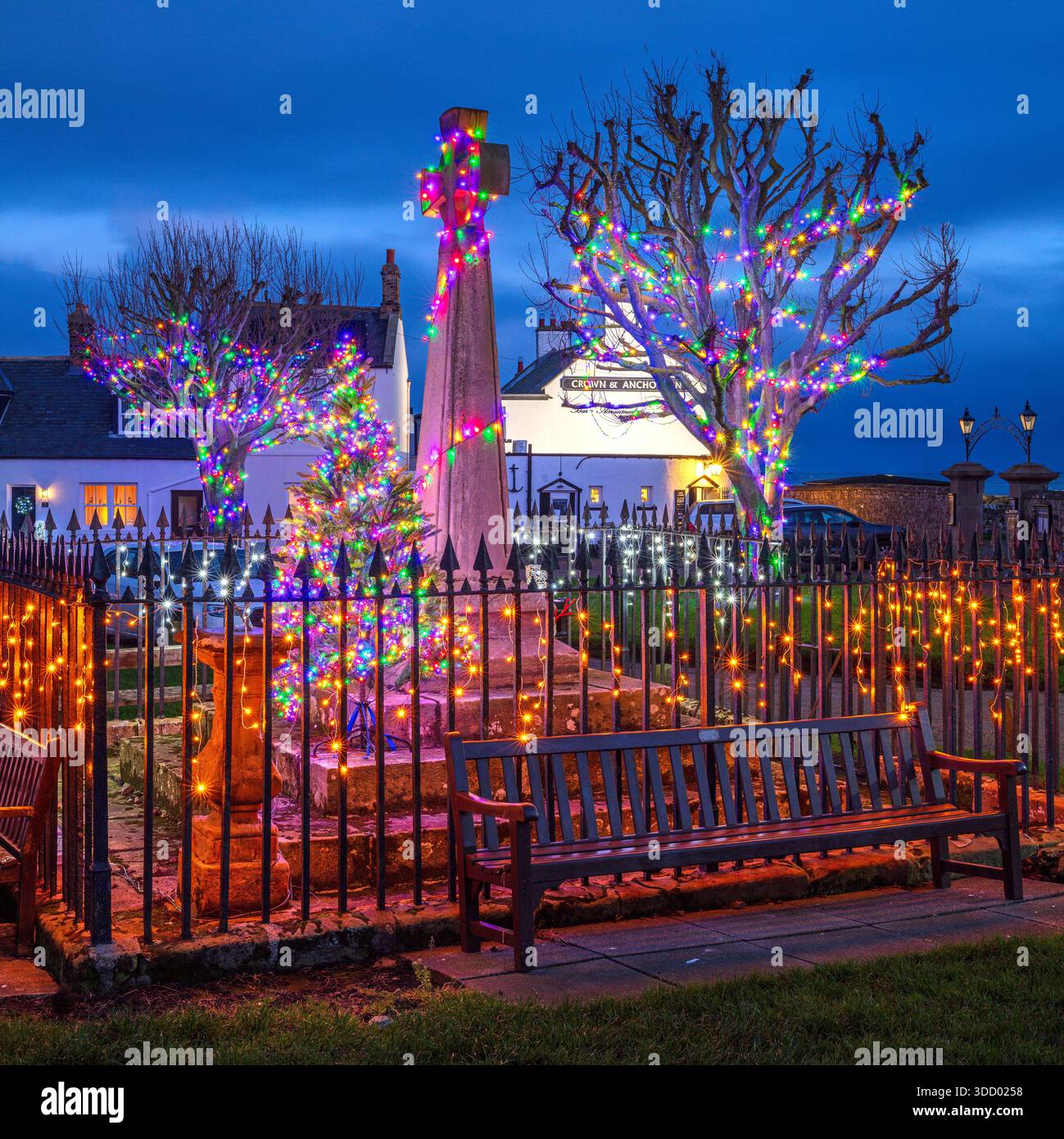 Una vista al crepuscolo di Natale dello spettacolo delle luci di Natale nella piazza del villaggio, memoriale di guerra sull'Isola Santa di Lindisfarne nel Northumberland Foto Stock