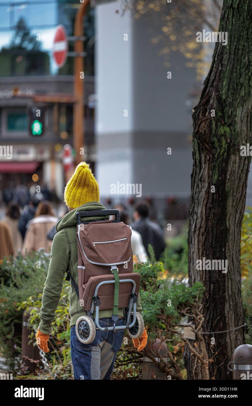 Un uomo in maglia gialla brillante porta lo zaino pieghevole sulla schiena mentre cammina lungo il marciapiede urbano tra pedoni e negozi. Foto Stock