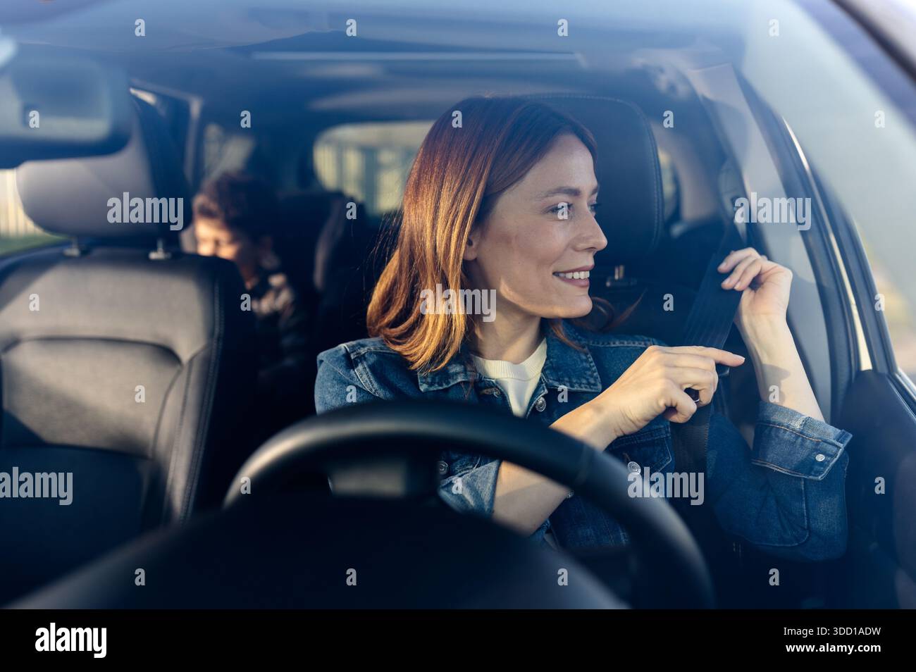 Una donna siede sul sedile di guida di un veicolo. Sorride quando allaccia la cintura di sicurezza. Un bambino è seduto nella parte posteriore. Foto Stock