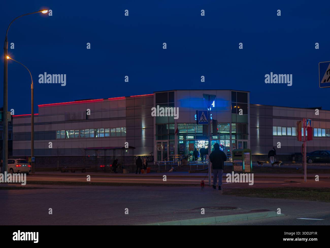La sera a Minsk mostra un basso centro commerciale con ingresso in vetro blu, LED rossi sul tetto, luci stradali calde, persone che entrano e escono, pedoni in un autobus Foto Stock