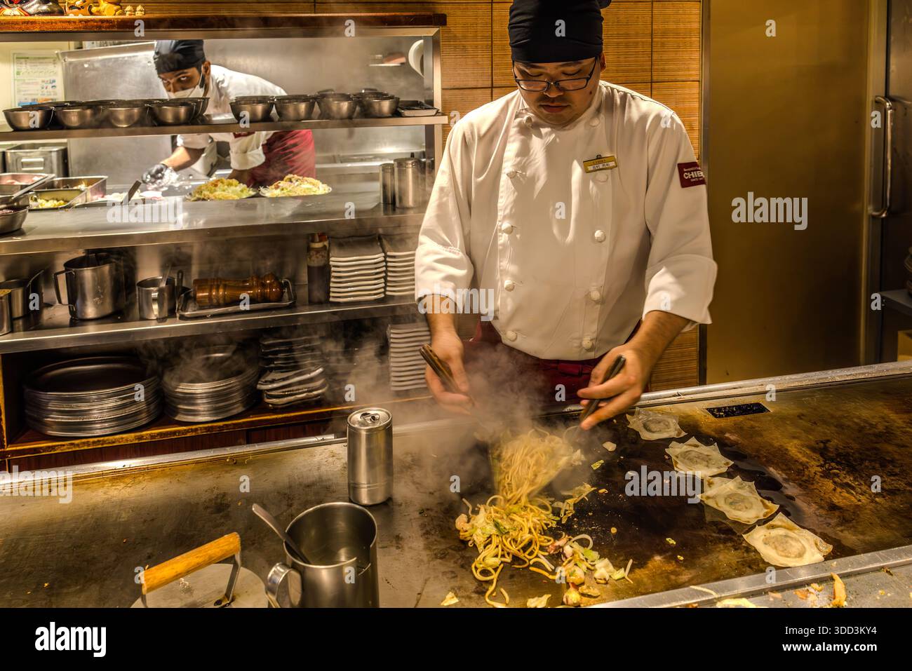 Uno chef frigge le noodles sulla griglia Teppan di un ristorante a Osaka, in Giappone, mentre un secondo chef prepara porzioni di okonomiyaki sullo sfondo. Il vapore sale dalle noodles e una lattina di bevanda è posata sul bordo della griglia. Il piatto è yakisoba – noodles di frumento fritti con cavolo e verdure – e si colloca accanto all'okonomiyaki tra i piatti alla griglia più popolari di Osaka. Foto Stock