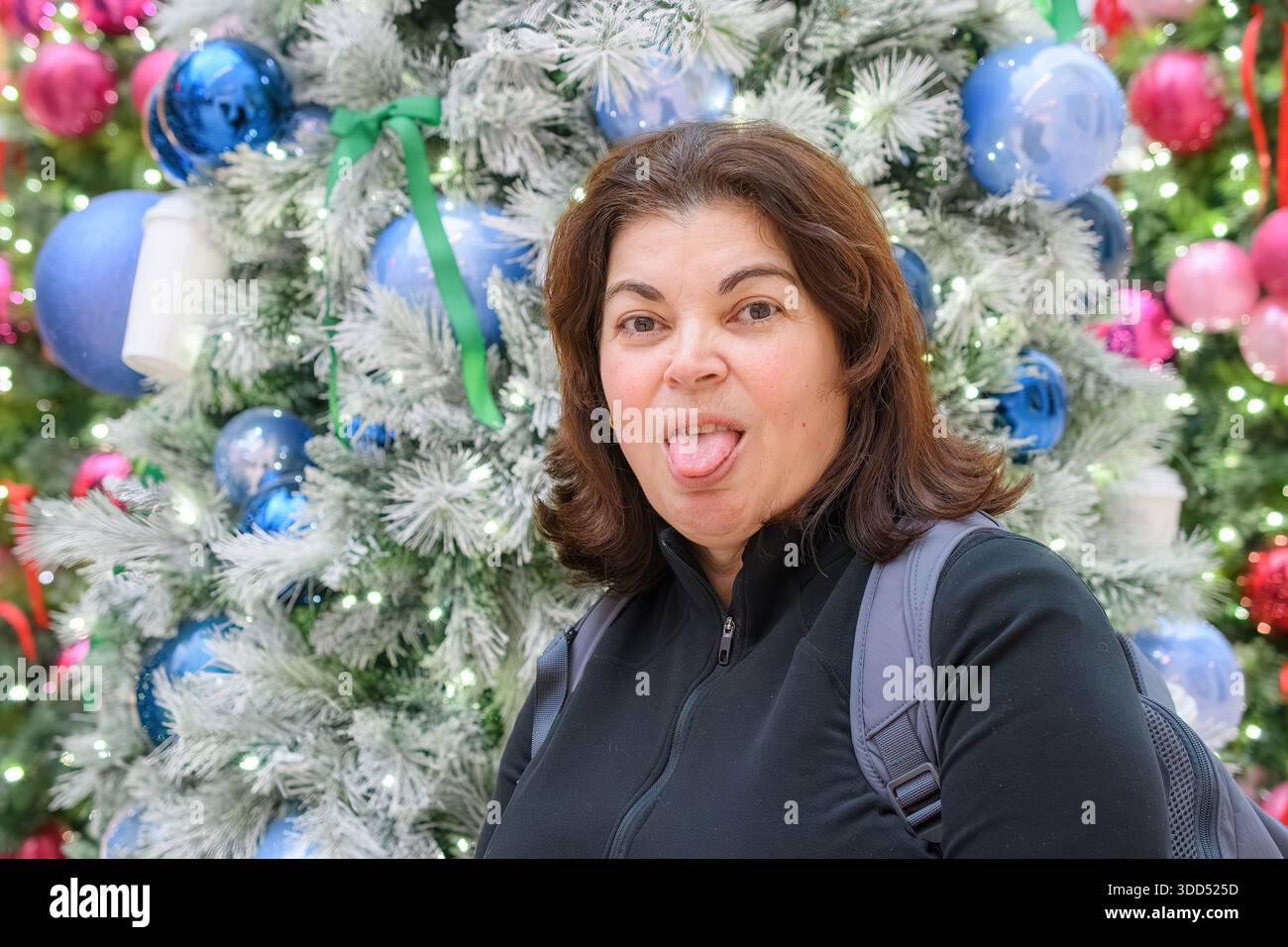 Donna giocosa che si attacca la lingua davanti a un albero di Natale decorato. Foto Stock