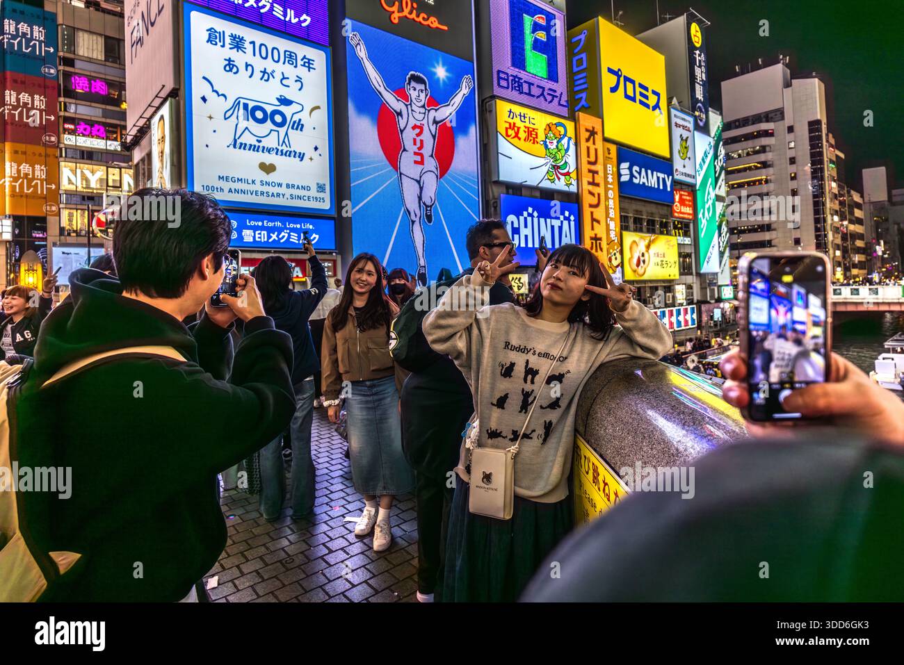 Una giovane donna si fa un selfie davanti ai cartelloni illuminati a Osaka, Giappone. L'uomo Glico raffigurato originariamente pubblicizza una barretta energetica contenente ingredienti di ostriche, prefettura di Osaka, Giappone Foto Stock