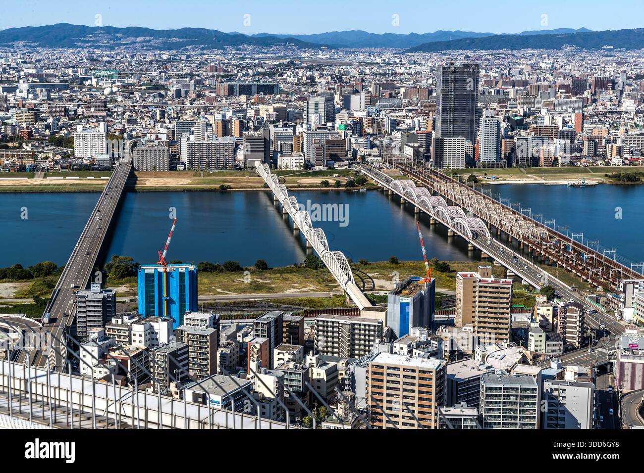 Diversi ponti attraversano un fiume nel paesaggio urbano densamente costruito di Osaka, Giappone, visti dall'Umeda Sky Building. Le gru di costruzione segnano progetti edilizi in corso lungo la riva; la vista dall'"Osservatorio del Giardino Fluttuante" a forma di anello del palazzo di 173 metri, completato nel 1993 da Hiroshi Hara, raggiunge le montagne. Foto Stock