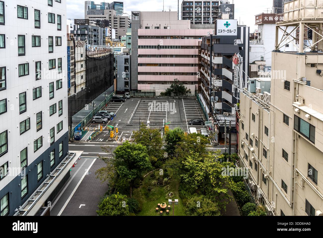 Un piccolo parco verde con alberi è incastrato tra un parcheggio e edifici residenziali e uffici a più piani. Osaka, Giappone. Foto Stock