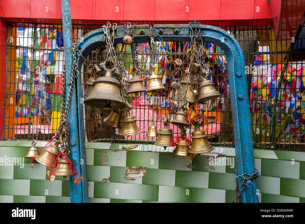 Diverse campane di preghiera in ottone pendono da una cornice in metallo blu intemprato di fronte a una parete verde a scacchi al Tempio Mahakal, Darjeeling. Le vivaci bandiere di preghiera e i dettagli architettonici rossi creano un ambiente vivace e spirituale. Foto Stock