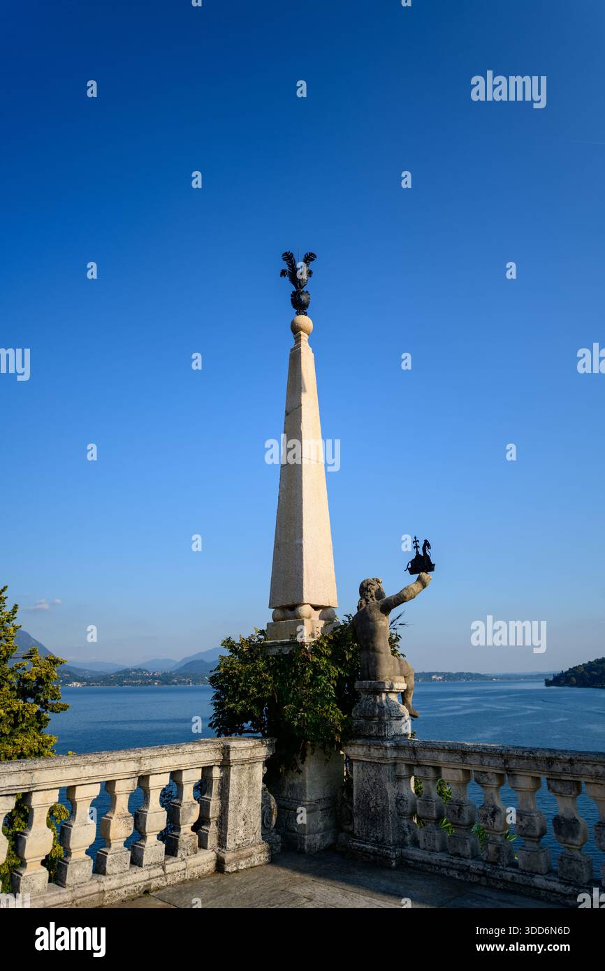 Un alto obelisco in pietra sormontato da ornati lavori in ferro sorge accanto a una statua classica su una terrazza con balaustra che si affaccia sulle acque blu del Lago maggiore. La luce fresca del sole mette in risalto l'architettura storica del giardino e la vista panoramica del lago nei Giardini Borromei, Italia. Foto Stock