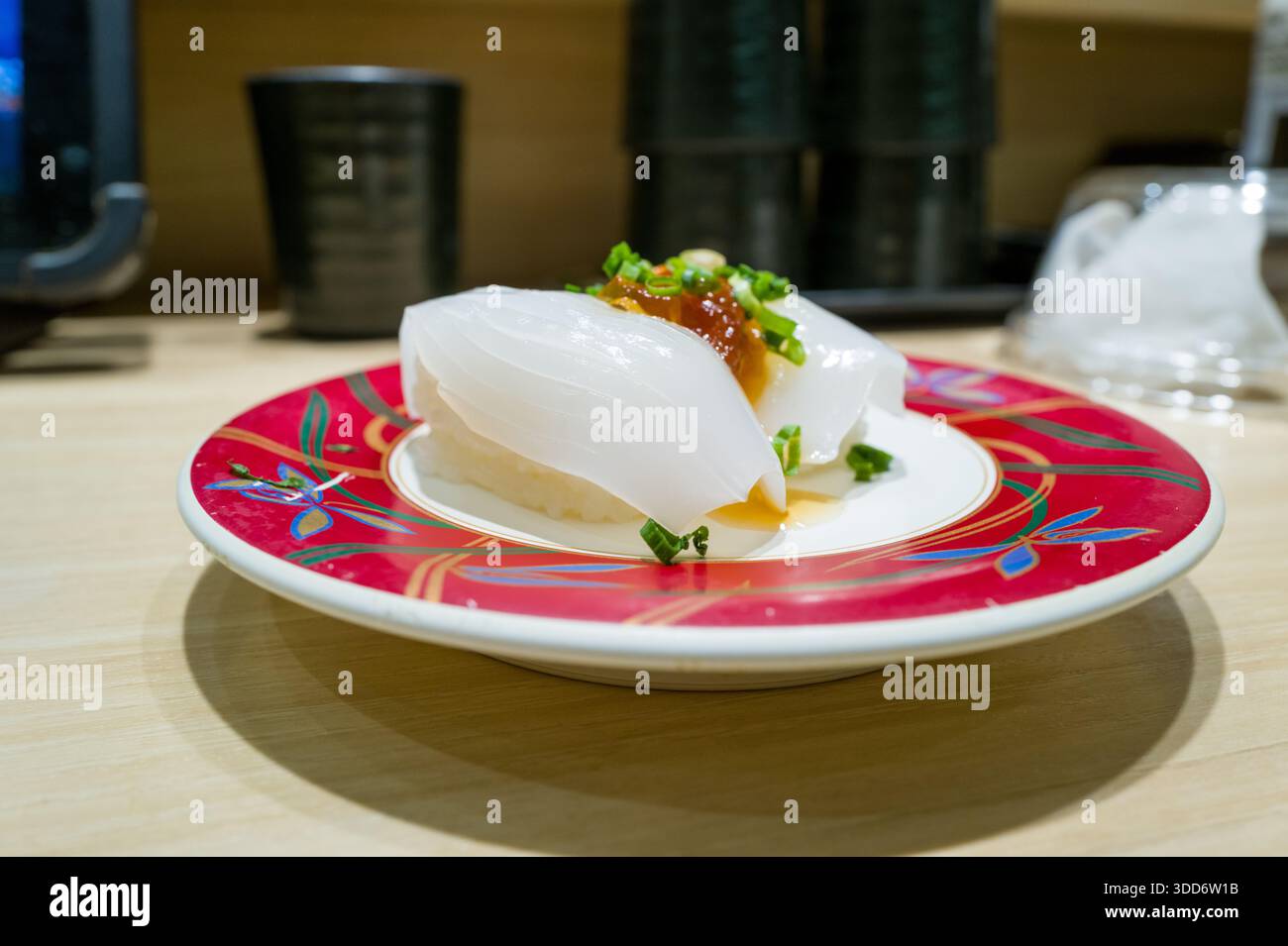 Due pezzi di sushi nigiri di calamaro traslucido conditi con uova di pesce d'arancia e cipolla verde tritata sono serviti su un piatto rosso vivace in cima ad un bancone di legno chiaro in un ristorante giapponese. L'illuminazione soffusa per interni mette in risalto la texture lucida e la presentazione fresca. Foto Stock