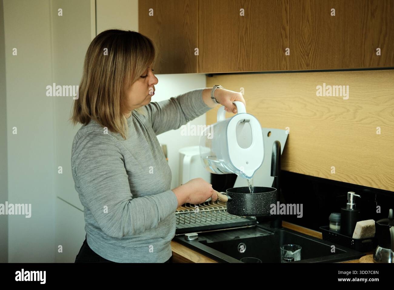 La donna sta in cucina versando acqua filtrata dalla caraffa nel piccolo recipiente sopra il lavandino. Filtraggio dell'acqua, vita sostenibile, acqua potabile pulita, eco Foto Stock