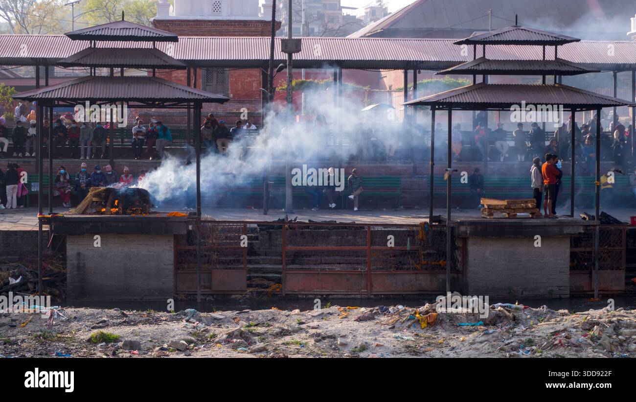 Le persone osservano rituali di cremazione indù su una riva del fiume mentre il fumo sale dalle pire funerarie, creando un'atmosfera solenne e spirituale radicata nel tradimento Foto Stock