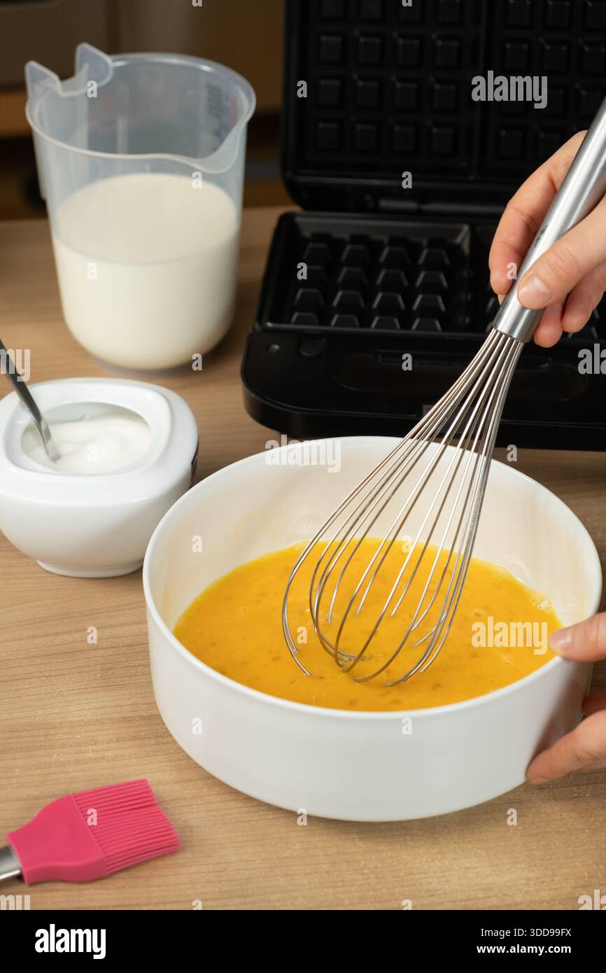 Usare una frusta per mescolare le uova crude in un recipiente bianco per cuocere i waffle Foto Stock