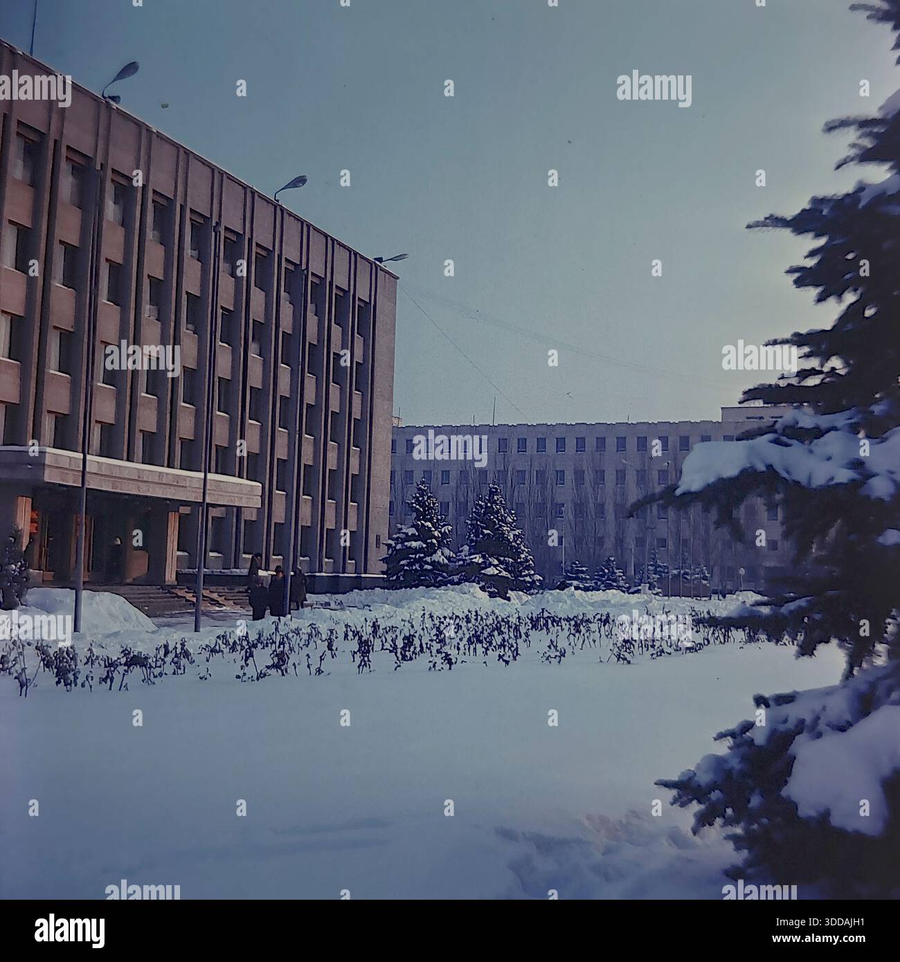 Slavyansk, Ucraina, URSS, anni 1980 Un paesaggio invernale soleggiato sulla centrale Piazza della Rivoluzione d'ottobre (ora Piazza Soborna). L'edificio grigio a più piani del Comitato Esecutivo della città (Gorispolkom) si erge dietro alberi di abete blu ricoperti di neve. Le profonde nevicate coprono le aiuole e i marciapiedi. Le persone in inverno sono visibili vicino all'ingresso principale. La foto cattura la grave bellezza di una fredda giornata invernale e la tipica architettura amministrativa sovietica. Foto Stock
