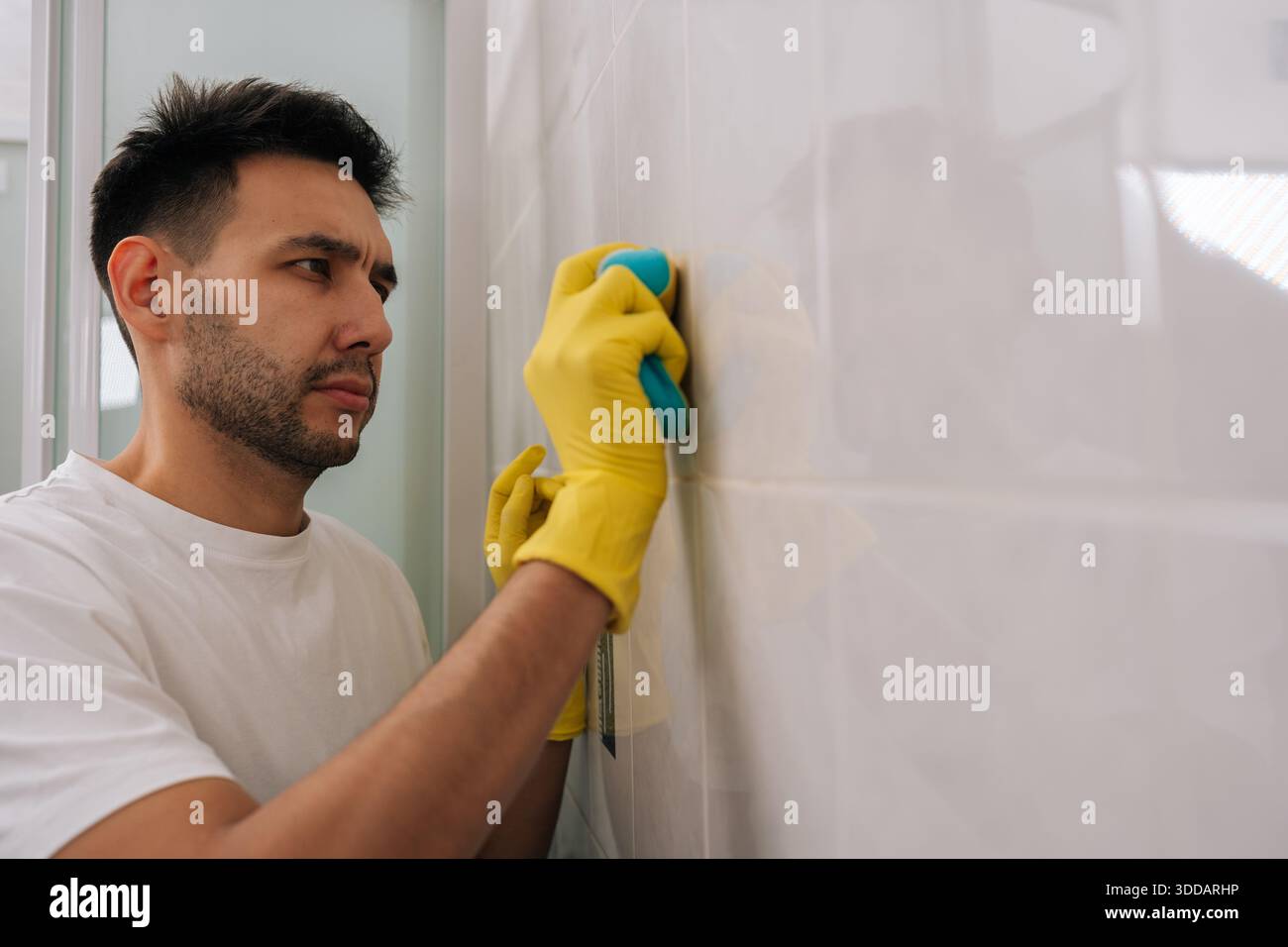 Uomo con barba concentrata che pulisce le piastrelle del bagno con spugna blu indossando guanti in gomma gialla, concentrandosi sulla pulizia delle pareti piastrellate bianche per Foto Stock