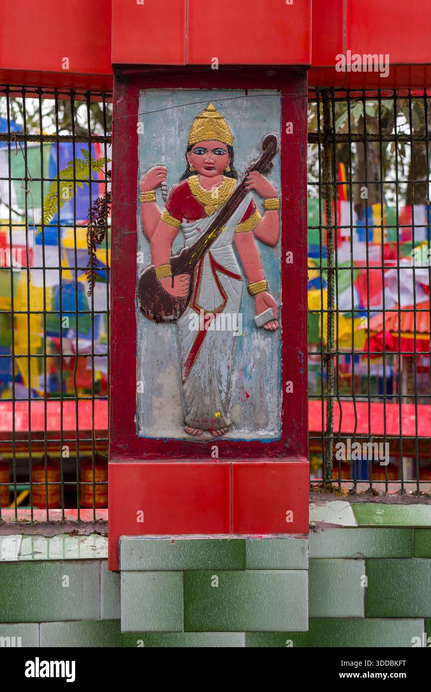 Colorata scultura in rilievo di una dea indù che tiene in mano uno strumento a corde incastonato in un muro rosso del santuario presso il Tempio Mahakal di Darjeeling. Bandiere di preghiera luminose e vegetazione forniscono uno sfondo vivido e spirituale. Foto Stock
