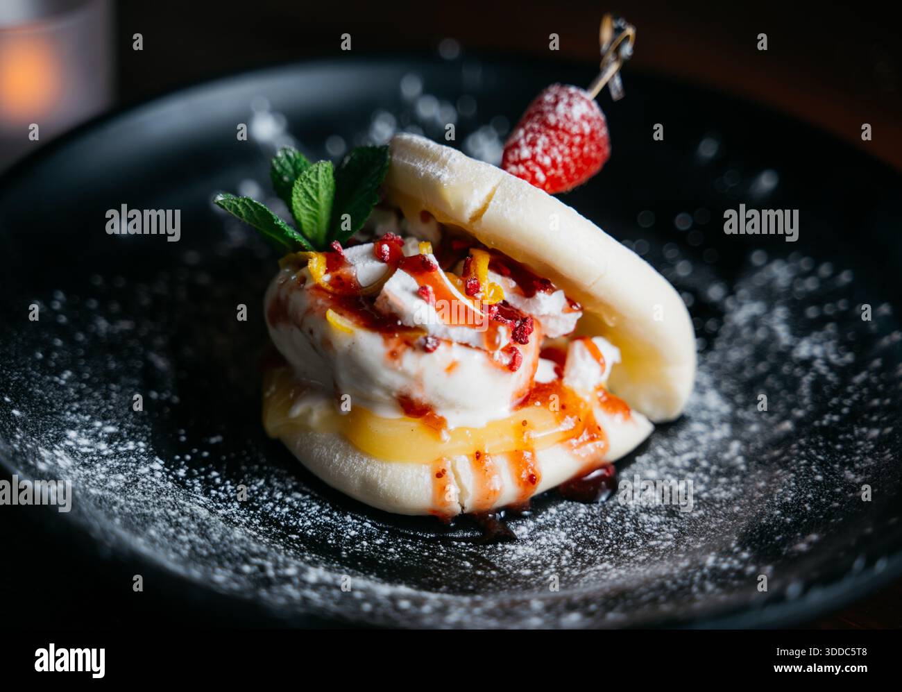 Un dessert con una fragola in cima e una foglia verde a lato. Il dessert è servito su un piatto nero Foto Stock