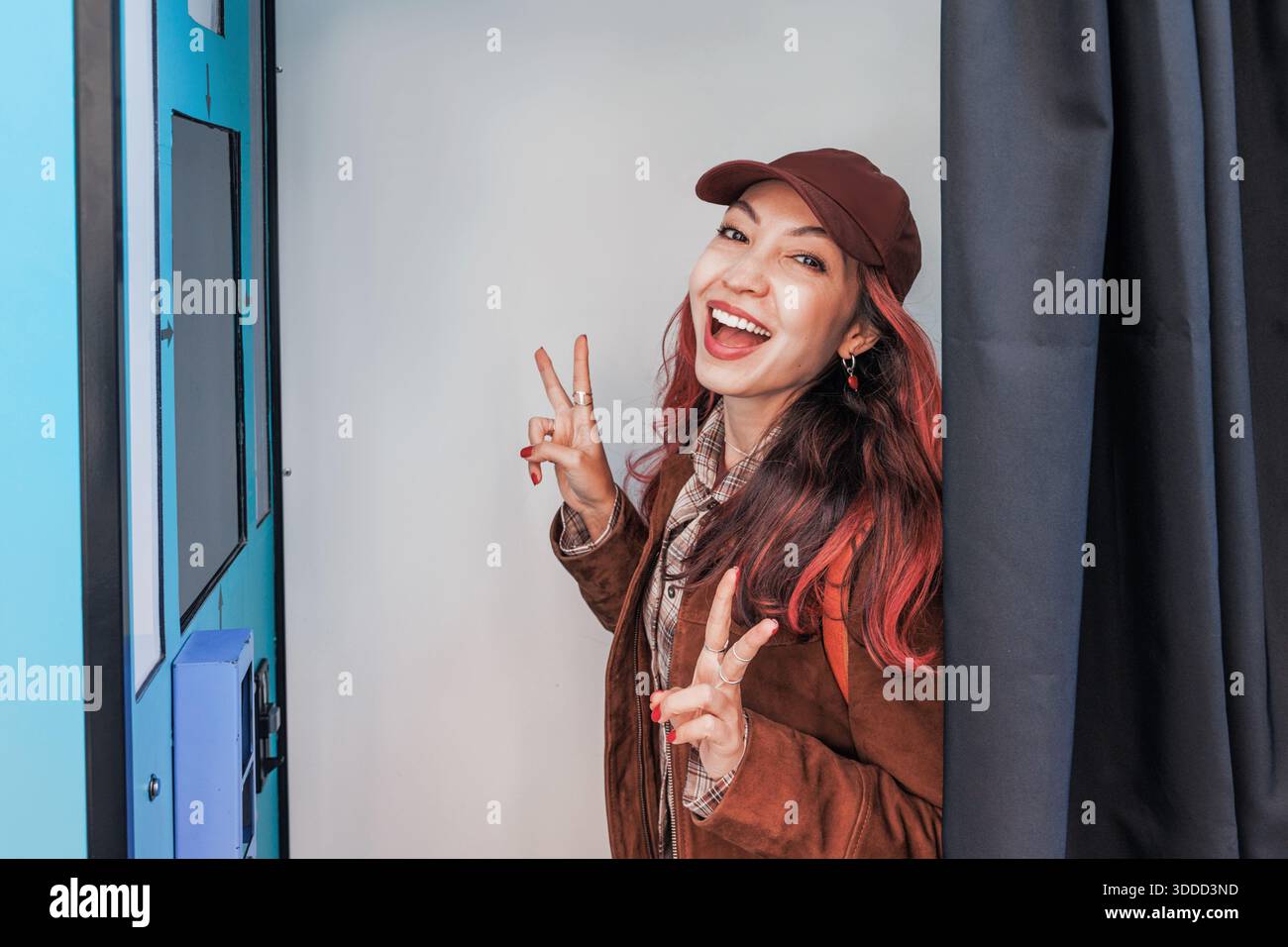 Giovane donna sorridente, che fa segnali di pace e mostra un'espressione vibrante all'interno di uno stand fotografico per ricordi divertenti Foto Stock
