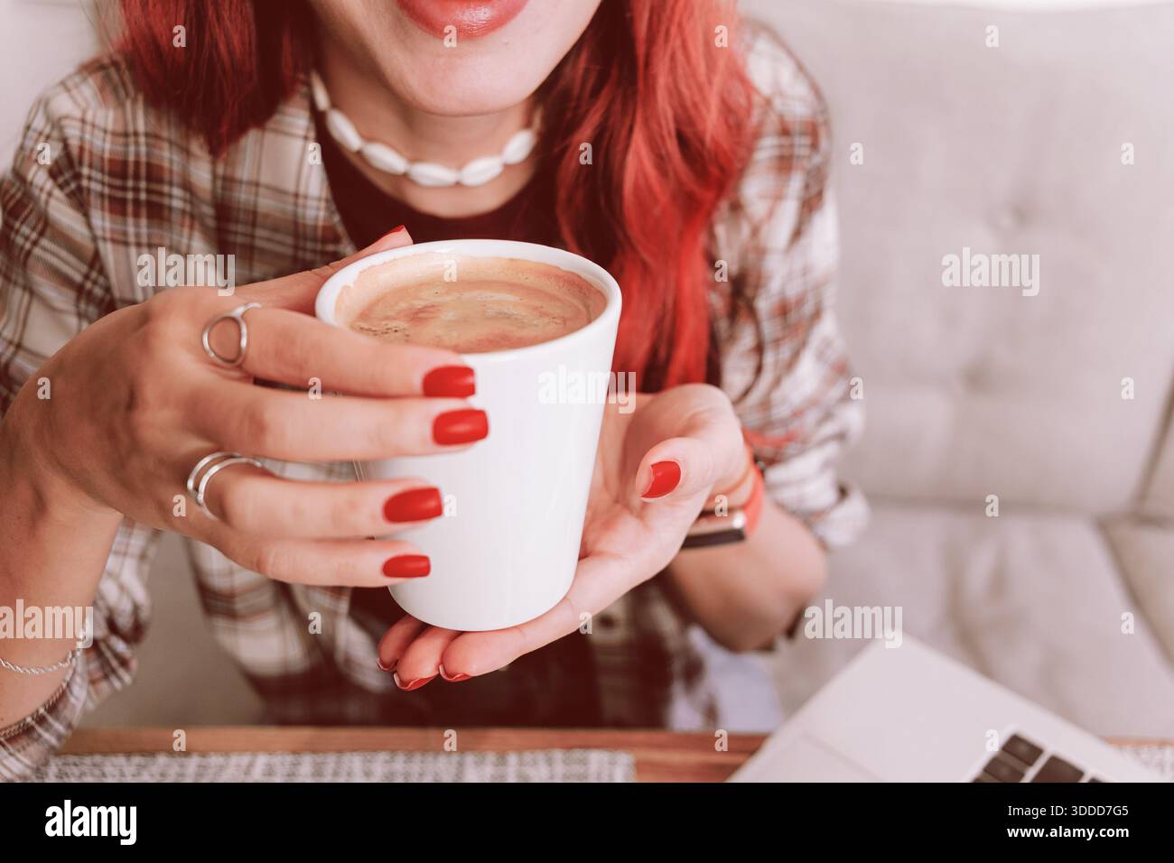 Donna dai capelli rossi con manicure rossa che tiene una tazza da caffè bianca calda vicino al computer portatile e si rilassa in un'atmosfera accogliente quando si lavora da casa Foto Stock