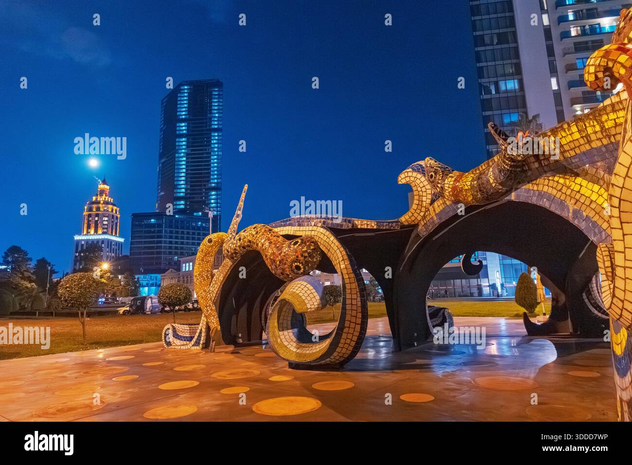 5 novembre 2025, Batumi, Georgia: Vibrante scultura di polpo a mosaico illumina la notte accanto ai moderni grattacieli e alla luna piena Foto Stock
