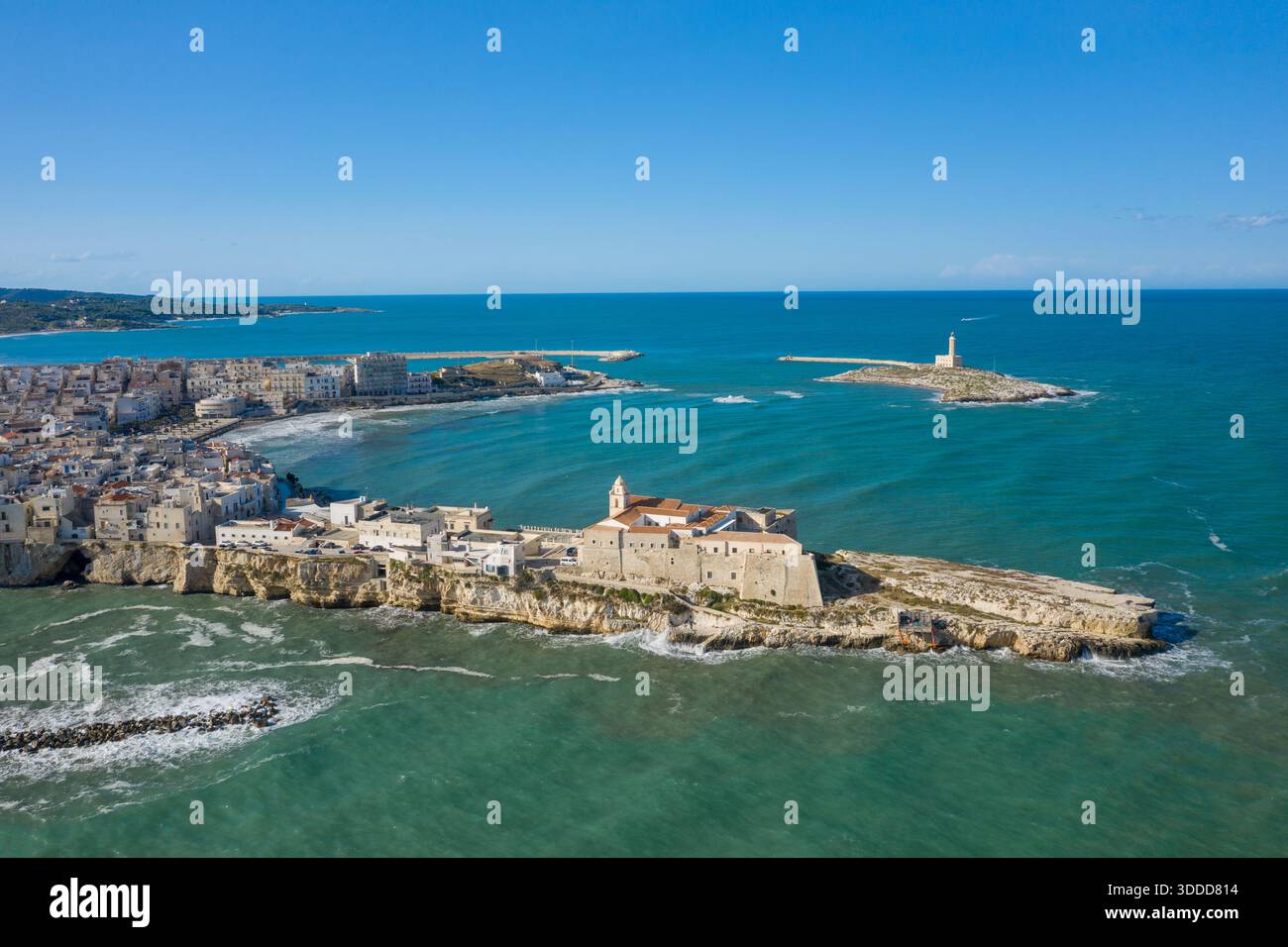 Una prospettiva aerea panoramica cattura la storica città vecchia di Vieste, in Italia, arroccata su aspre scogliere calcaree con il faro di Santa Eufemia in piedi su una piccola isola al largo. Il mare Adriatico turchese e l'ampio cielo blu fanno da cornice a questo iconico paesaggio costiero mediterraneo. Foto Stock