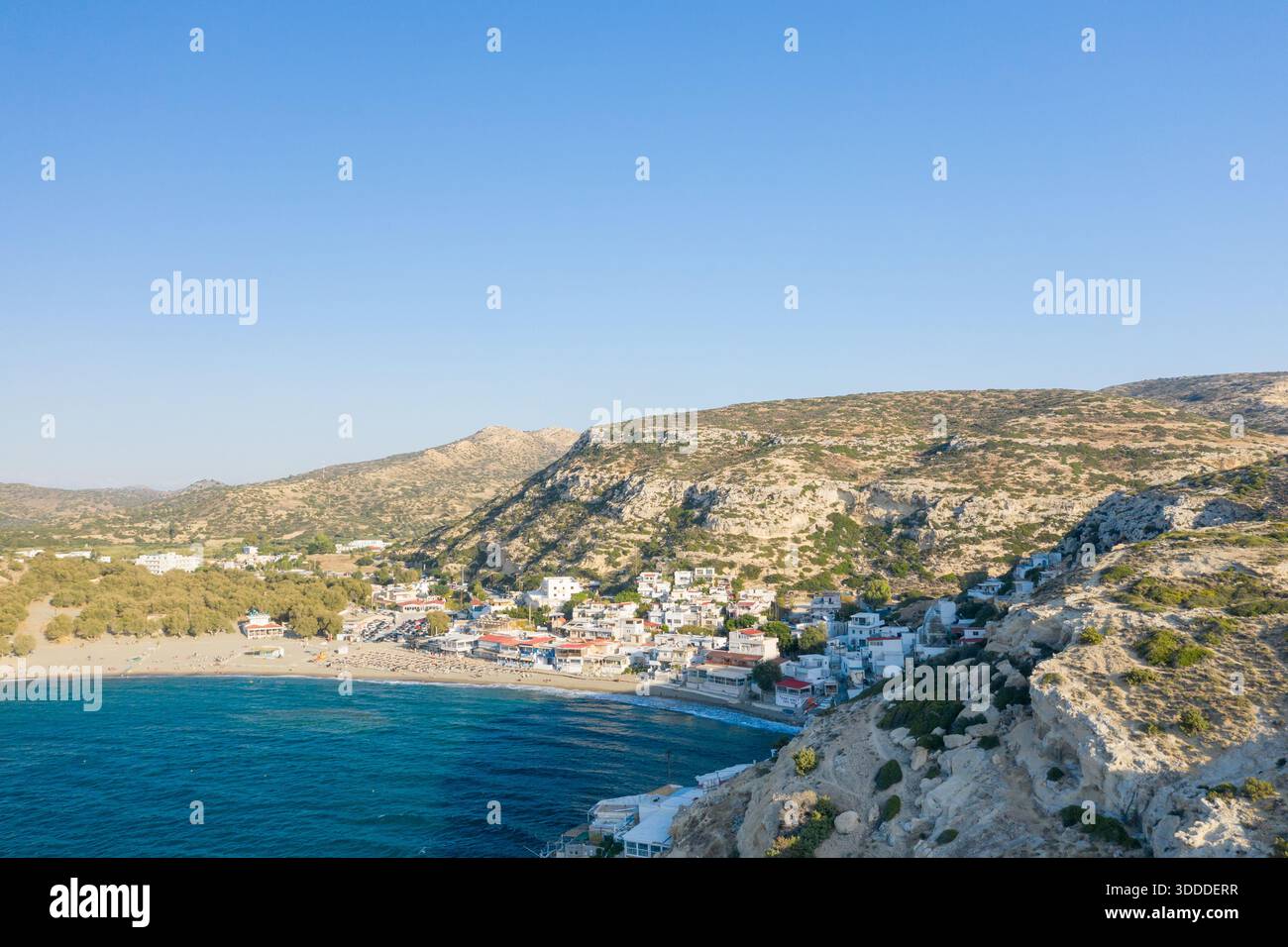 Il villaggio di Matala sorge tra una baia sabbiosa e colline calcaree illuminate dal sole sulla costa meridionale di Creta. L'acqua blu cristallina contrasta con le formazioni rocciose testurizzate e gli edifici imbiancati sotto un cielo senza nuvole. Foto Stock