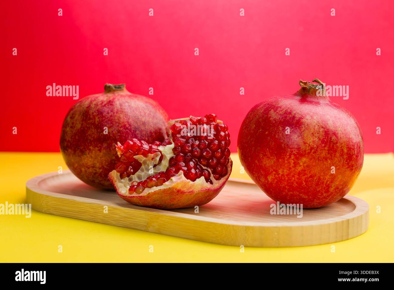 Due melograni interi e uno tagliato aperto con semi rossi sono su un vassoio di legno Lo sfondo ha colori rosso e giallo luminosi. Questa scena si svolge io Foto Stock