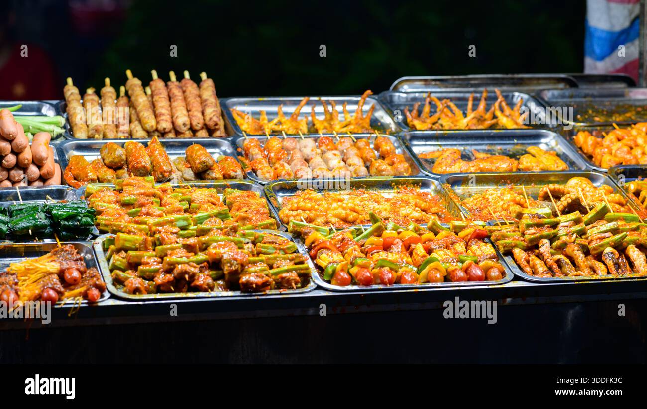 Un vivace assortimento di spiedini marinati, salsicce e verdure alla griglia è disposto su vassoi di metallo in un vivace mercato notturno di CAN Tho, Delta del Mekong, Vietnam meridionale. La scena è illuminata da luci luminose, che mettono in risalto le salse colorate e la varietà di cibo di strada. Foto Stock