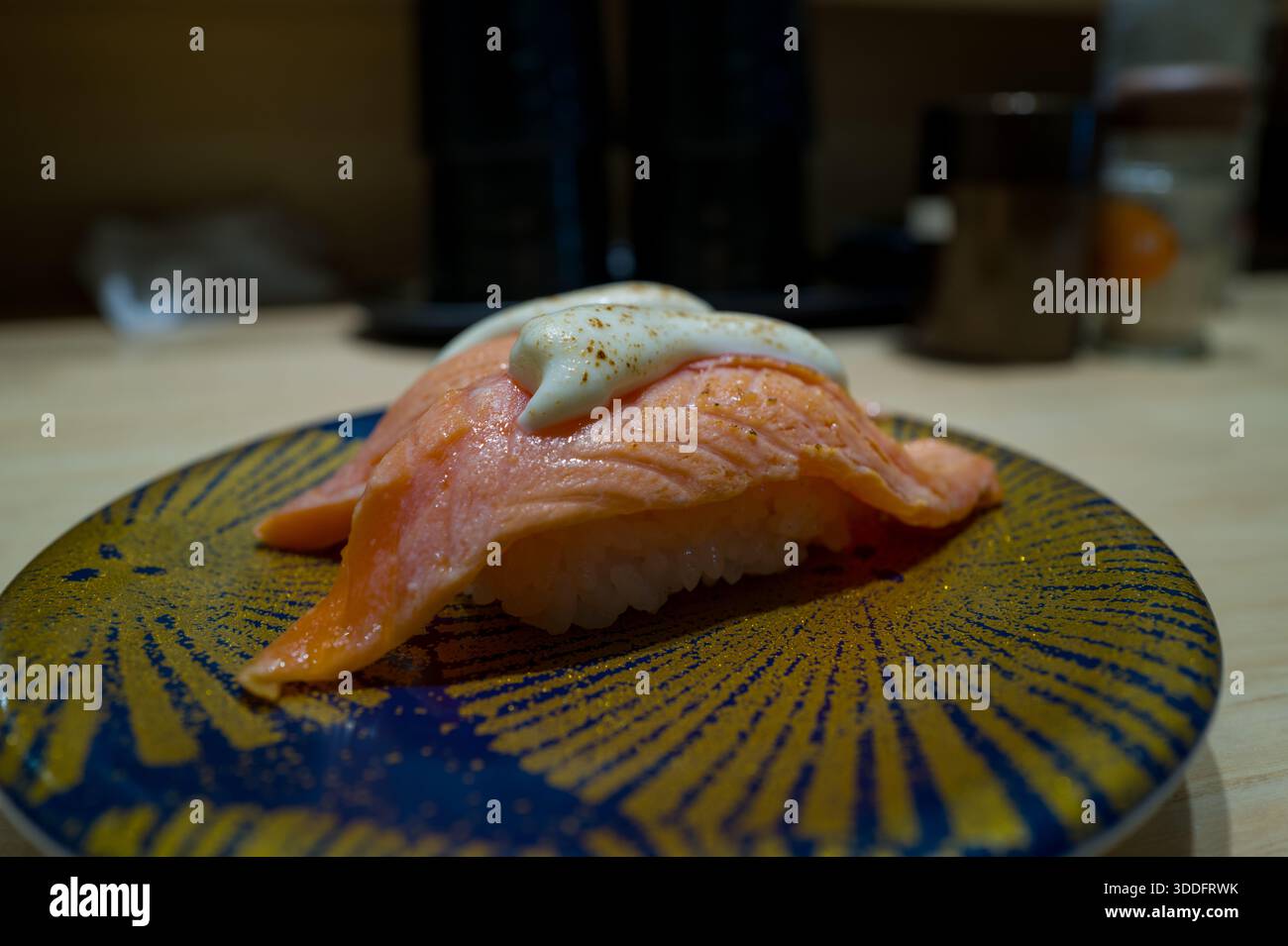 Un pezzo di sushi di salmone condito con un cucchiaio di cremosa salsa bianca è presentato su un piatto decorativo blu e oro, collocato su un bancone di legno chiaro in un interno del ristorante delicatamente illuminato. Foto Stock