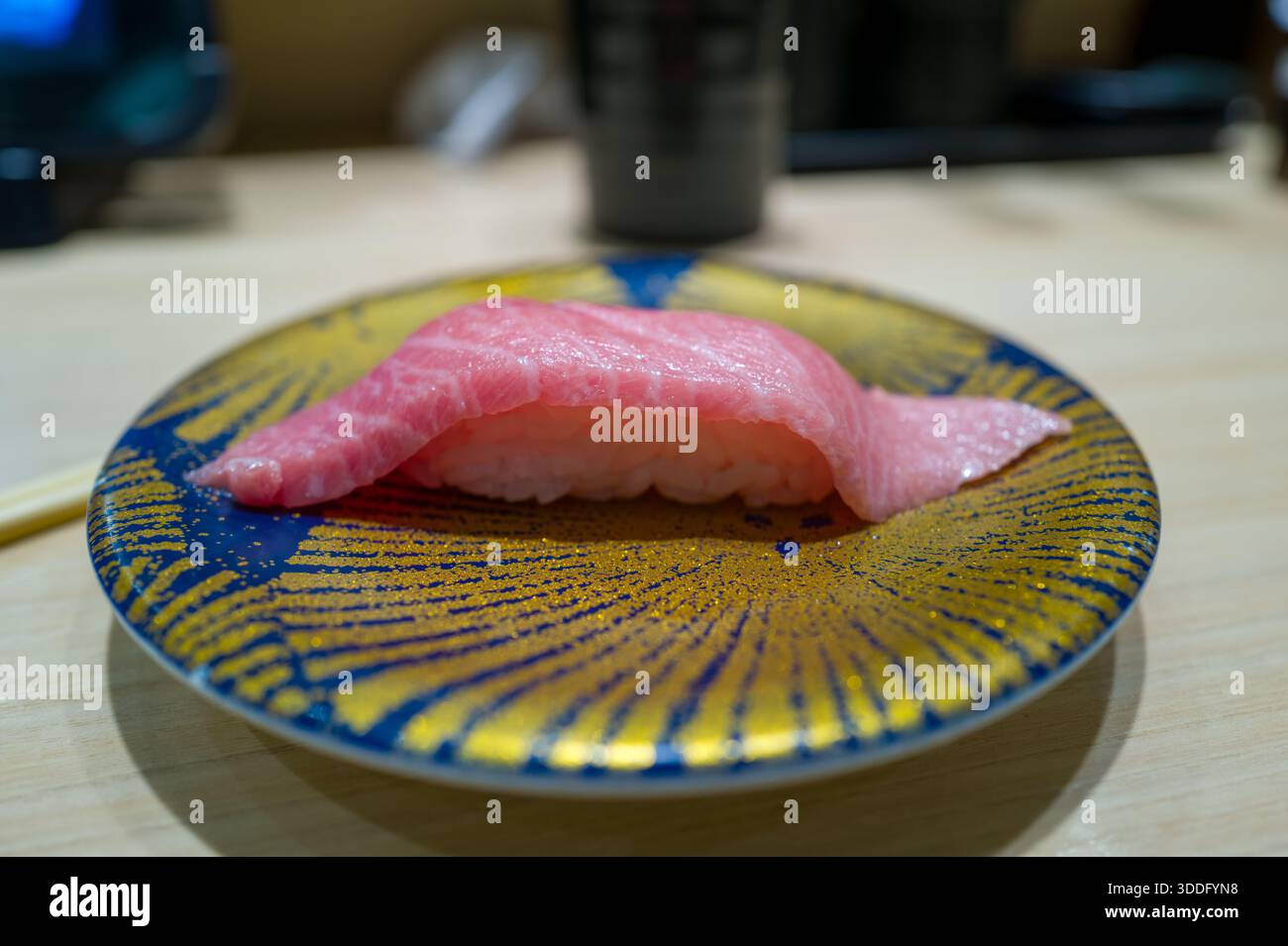 Un pezzo unico di otoro nigiri sushi con tonno rosa marmorizzato servito su un piatto blu e dorato vivace. Il sushi e' presentato su un banco di legno chiaro in un ristorante Giapponese con luce soffusa. Foto Stock