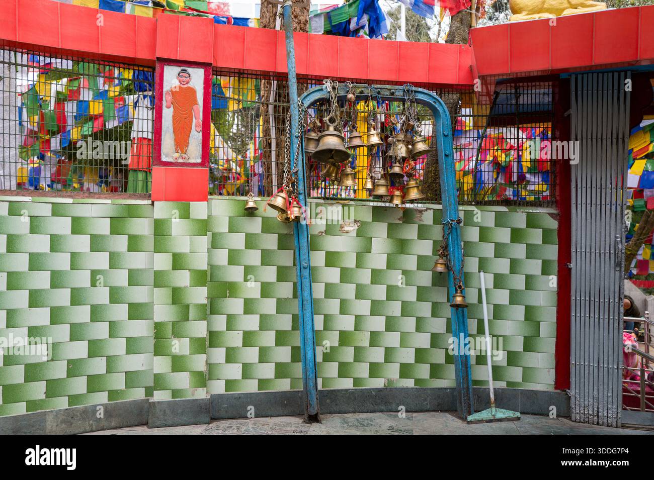 Un gruppo di campane in ottone appese su una struttura in metallo blu contro una parete piastrellata verde al Tempio Mahakal, Darjeeling. Bandiere di preghiera multicolore e una figura dipinta aggiungono dettagli vivaci e spirituali. Foto Stock