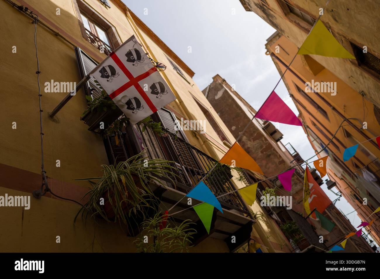 Bandiera sarda esposta su un balcone con piante in vaso sopra una stretta strada di Bosa, in Italia. I vivaci striscioni a pennant aggiungono un colore festoso alle storiche facciate gialle e arancioni sotto la luce soffusa del giorno. Foto Stock