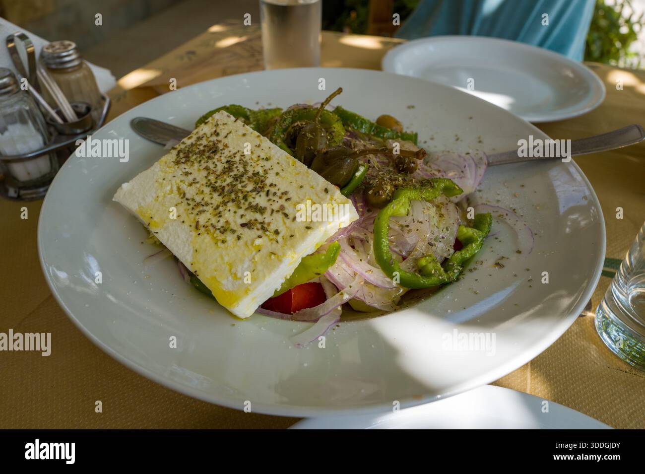 Insalata greca fresca servita in una ciotola bianca, con una fetta spessa di feta, peperoni verdi, cipolle rosse, olive e un filo di olio d'oliva, bagnata dalla luce naturale su un tavolo rustico. Foto Stock
