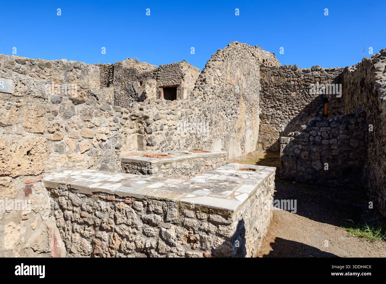 Le mura di pietra vulcanica intemperie racchiudono uno storico banco romano con inserti in marmo nei quartieri archeologici di Pompei, Italia. La luce del sole e il cielo azzurro esaltano la struttura delle rovine e dello spazio aperto. Foto Stock