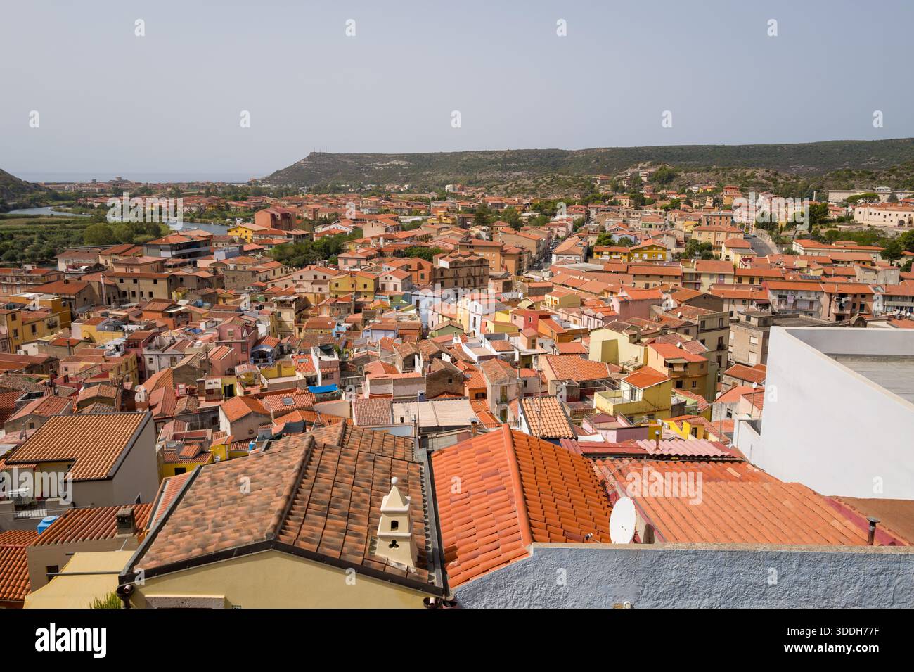 Ampia vista sui tetti in terracotta e sulle facciate pastello di Bosa, in Sardegna, con dolci colline verdi e il fiume in lontananza. La luce soffusa del giorno esalta i toni caldi e l'invitante fascino mediterraneo della città. Foto Stock