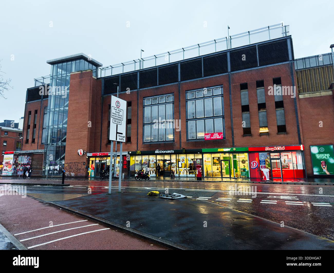 Manchester, Regno Unito - 06 dicembre 2025: Rainy Manchester Street con vetrine in mattoni, torre di vetro e negozi affollati. Foto Stock